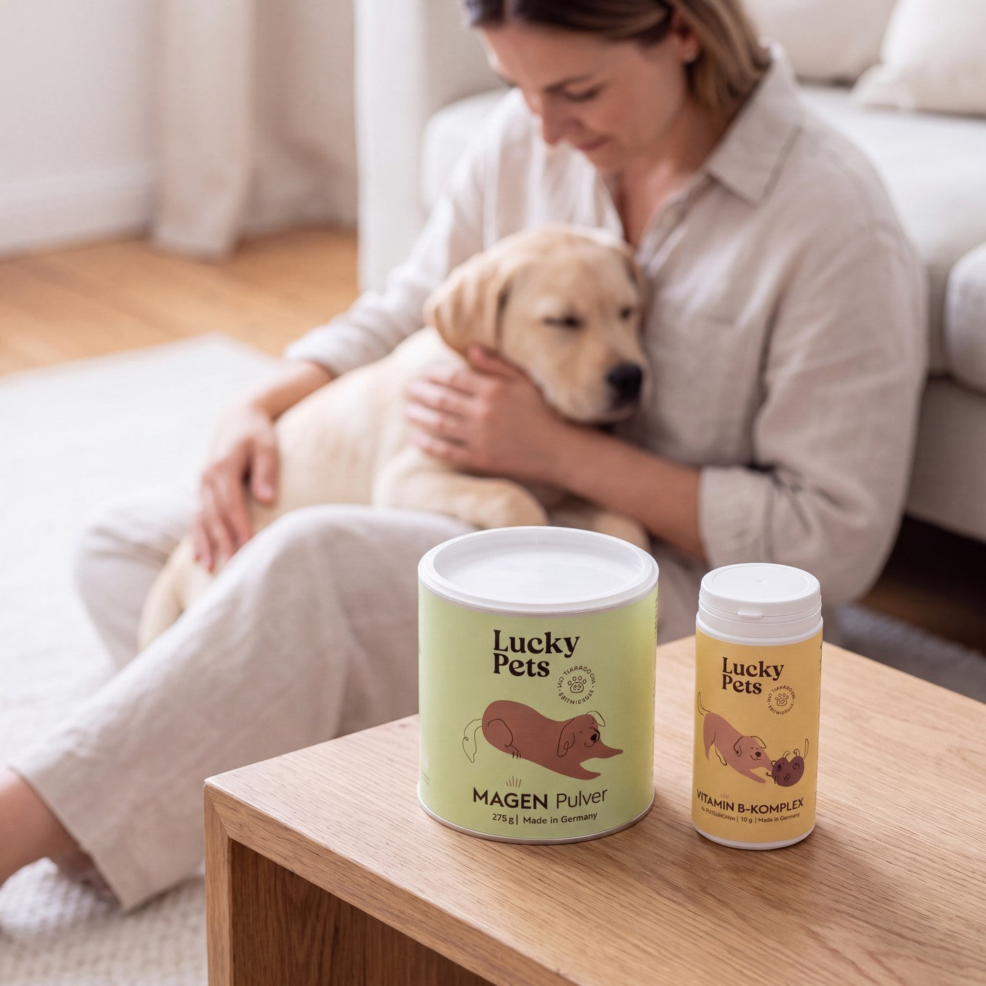 Frau sitzt im Wohnzimmer auf dem Boden mit ihrem schlafenden Labrador-Welpen auf dem Schoß, auf dem Couchtisch Magenpulver und Vitamin B-Komplex