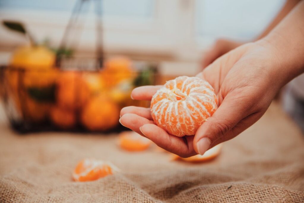 Eine geschälte Mandarine liegt in der Hand einer Person. Im Hintergrund sind noch einige Früchte zu sehen.